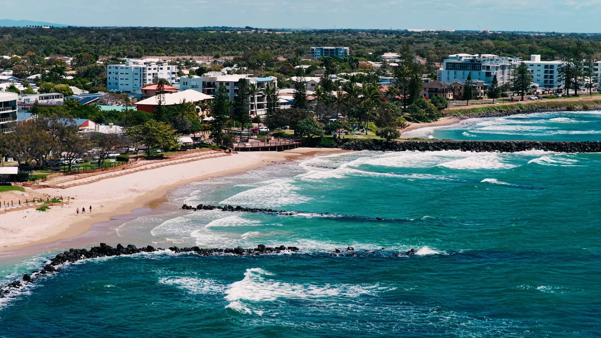 Aerial photograph of Kellys Beach, Bargara: the surf breaking over the rock groynes, the sand and the foreshore park, with the township's apartments and trees behind